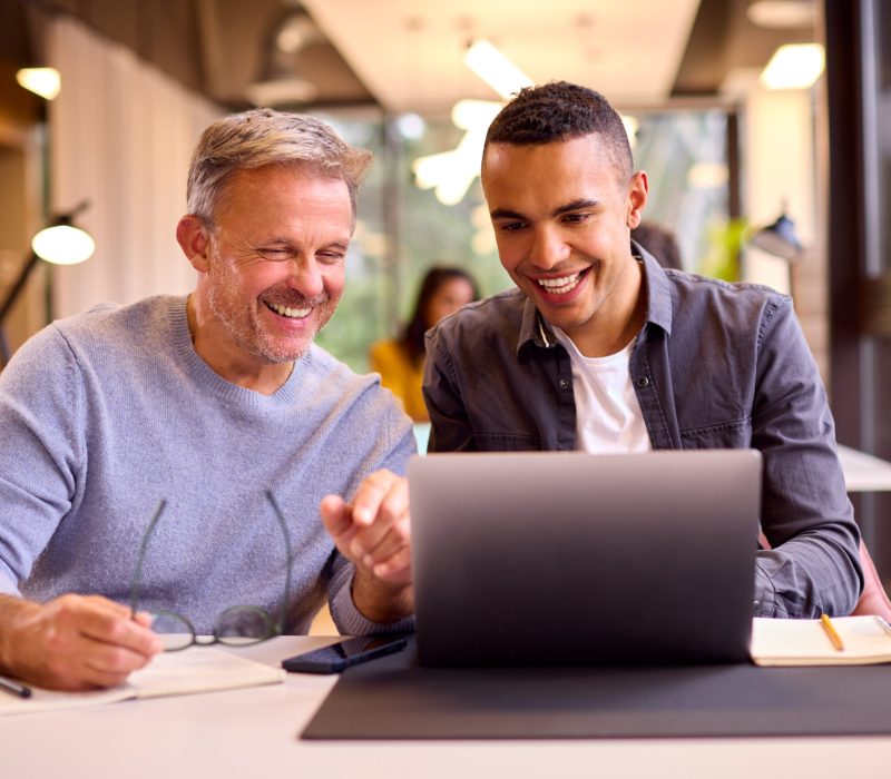 Mature Businessman Mentoring Younger Colleague Working On Laptop At Desk